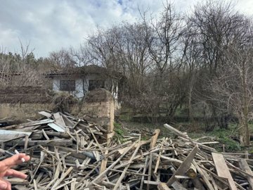 Garden area cluttered with wooden debris and rubble with the old house partially visible behind leafless trees.