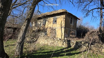 Exterior view of a two-storey rural house with wooden windows and a stone wall, surrounded by leafless trees under a clear blue sky.