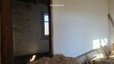 Interior view showing a white-walled room with a doorway opening to another space, sunlight casting shadows, and hay scattered on the floor.