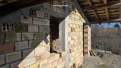 Partially constructed brick wall with window openings and visible wooden roof beams in unfinished building.