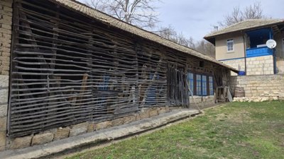 A traditional wooden fence with horizontal slats and a stone base next to a rustic house with blue window frames.