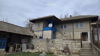 Side perspective of the Bulgarian house featuring a blue wooden balcony and stone stairs leading to the entrance