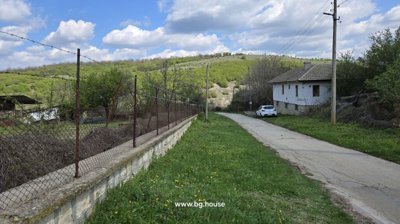 View along a village road bordered by a wire fence and a white house, surrounded by green hills and trees under a partly cloudy sky.