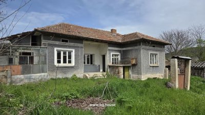 Front view of the Bulgarian rural house showing grey walls, tiled roof, and surrounding garden area