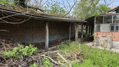 Brick outbuilding with wooden supports and glass porch adjacent to the house, surrounded by overgrown vegetation