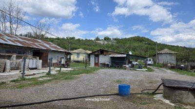 View of multiple farm buildings and a yard area with gravel and grass, surrounded by trees and hills.