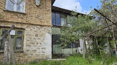 Side perspective of the two-storey house featuring stone and brick walls, an enclosed terrace, and surrounding green plants.