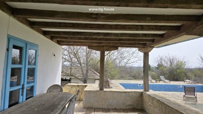Covered wooden veranda with dining table and chairs overlooking a swimming pool and garden area.