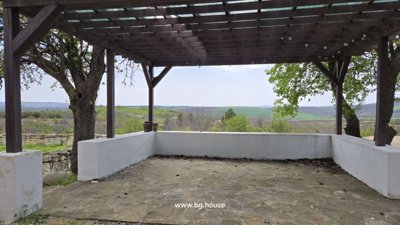 Stone-floored terrace with white walls and wooden pergola overlooking the Bulgarian countryside with trees.