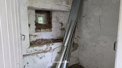 A small room showing whitewashed walls, a small window with bricks around it, and metal pipes leaning against the wall.