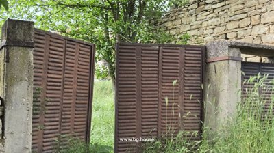 Close-up of a rusty metal gate next to a stone wall with green plants growing around it.