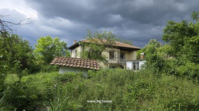 View of the yellow two-storey house with a balcony, surrounded by dense green vegetation and a cloudy sky.