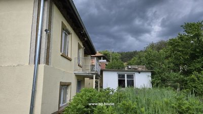 Yellow house with balcony and adjacent white outbuilding surrounded by tall grass and trees.
