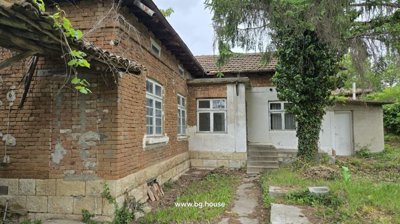 Exterior view of a traditional Bulgarian brick house with white window frames and a tiled roof, set in a garden with trees and plants.