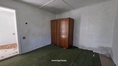 Interior view of a room with a green carpet, a wooden wardrobe, and white walls showing signs of wear and peeling paint.