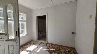 An empty indoor room featuring white walls, large windows letting in natural light, and dry leaves scattered across the floor.