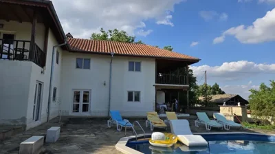 View of the white two-storey house with a tiled roof and a swimming pool surrounded by sun loungers on a stone patio.