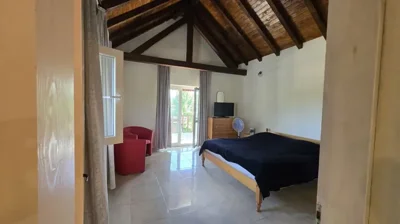 A bedroom featuring exposed wooden ceiling beams, a double bed with black bedding, a red armchair, and doors leading to a balcony.