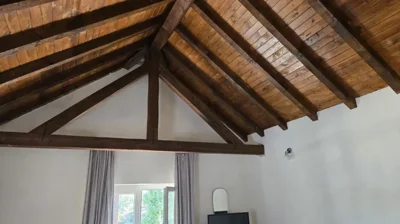Detailed view of the wooden ceiling beams above a window framed by light curtains in a bedroom.