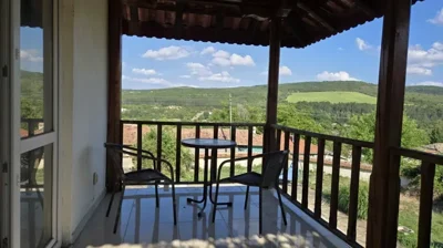 A covered balcony furnished with a small round table and two chairs overlooking green hills and rooftops.