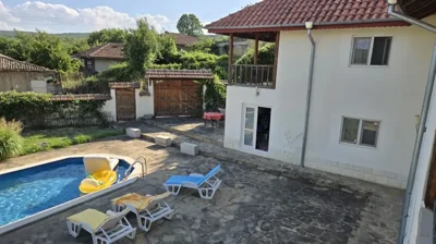 Stone patio area with swimming pool and white two-storey house featuring a wooden balcony and garden gate.