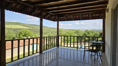Covered balcony with tiled floor, wooden railing, table and chairs offering views of green fields and hills.