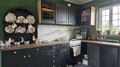Kitchen area showing black lower cabinets, wooden countertops, a stove, microwave, and a decorative dish rack with plates and cups against a green wall.