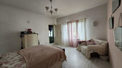Bedroom showing a double bed, a cream-colored dresser, and a window with sheer curtains allowing natural light to enter.