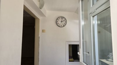 White painted wall with a round clock showing 12:05, a window with white frame, and a small open hatch below the clock.