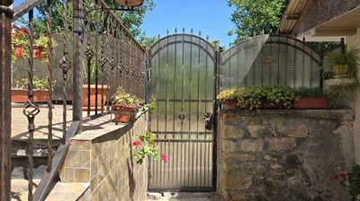 Outdoor view of a metal gate with decorative elements, stone wall, and flower pots with plants on the side.