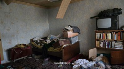 Interior room showing wallpapered walls, wooden furniture including a bookshelf, and various items scattered around.