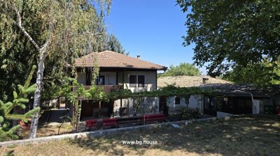View of the two-storey Bulgarian house with a tiled roof, shaded by mature trees and featuring a garden area with a grapevine trellis.