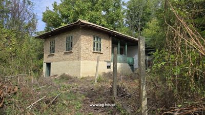 Side perspective of a traditional Bulgarian house featuring a stone cellar, wooden balcony with green railings, and garden area with trees and shrubs.