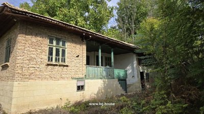 Traditional Bulgarian house showing wooden balcony with green railings and stone ground floor, surrounded by dense greenery and garden plants.