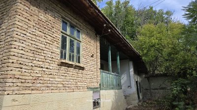 Close-up view of a traditional Bulgarian house featuring a wooden balcony with green railings and stone lower walls, surrounded by trees and bushes.