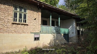 Traditional Bulgarian house showing wooden balcony with green railings and stone ground floor illuminated by sunlight, surrounded by garden plants.