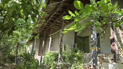 View of a traditional Bulgarian house facade featuring a wooden balcony, green doors, and leafy trees.