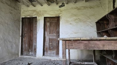 A rustic interior room featuring two wooden doors, rough plaster walls, exposed ceiling beams, and an old wooden table with shelves.