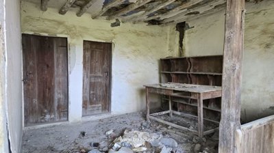 Wide angle of a rustic interior room with two wooden doors, exposed ceiling beams, an old wooden table, and debris on the floor.