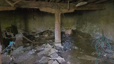 A large rustic cellar featuring stone and plaster walls, wooden beams, a central wooden post, and many glass jars and debris scattered on the floor.