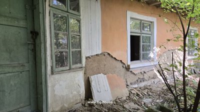 Detailed view of the house wall showing peeling plaster, a green wooden door, and windows surrounded by vegetation.