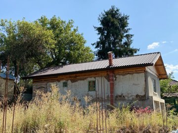 Exterior view of an old Bulgarian house featuring a newly tiled roof and chimney, with overgrown vegetation in the foreground.