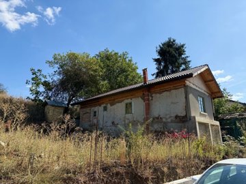 Side view of an old Bulgarian house showing a new roof and chimney, surrounded by dry garden plants and trees.