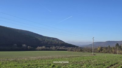 Wide open green field bordered by wooded hills under a clear blue sky with visible airplane contrails.