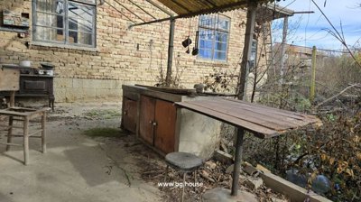Patio area with wooden table, stool, and hanging kitchen utensils under a translucent roof beside brick walls.