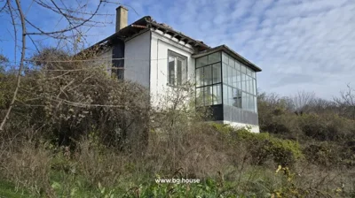 Exterior view of a white two-storey house with a glass-enclosed balcony and overgrown bushes around it