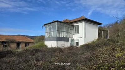 White two-storey house featuring a glassed porch and tiled roof, surrounded by wild bushes