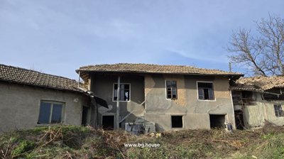 Exterior view of an old Bulgarian house with tiled roof and several windows, showing the rustic facade and surrounding dry vegetation.