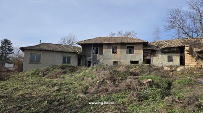Wide exterior view of an old Bulgarian house with tiled roof and several windows, surrounded by dry vegetation and garden space.
