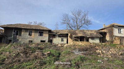 Wide exterior view of an old Bulgarian house with tiled roof and several windows, surrounded by dry vegetation and garden space.